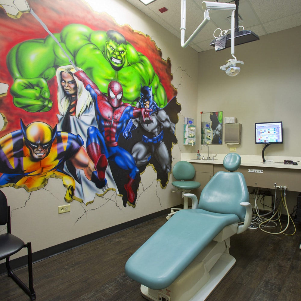 Dental operatory with Marvel superhero mural, treatment chair, and dental equipment for pediatric patients.