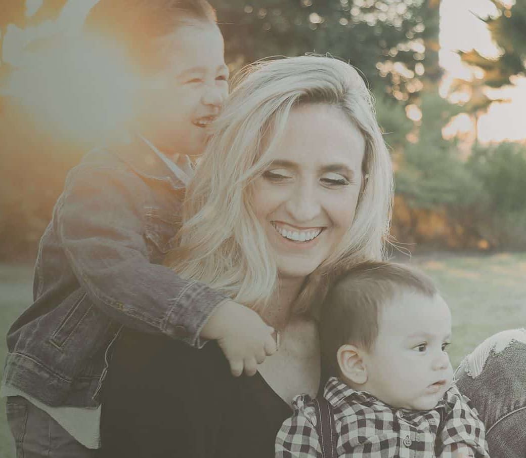Woman and two young children smiling outdoors during golden hour sunset.
