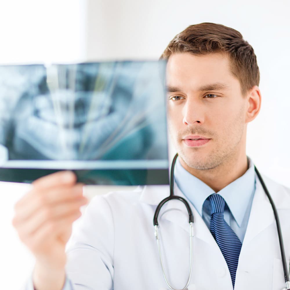Male dentist in white coat examining dental X-ray against light background with stethoscope.