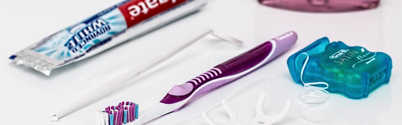 Dental care products including toothpaste tube, purple toothbrush, small brush, and turquoise container on white surface.
