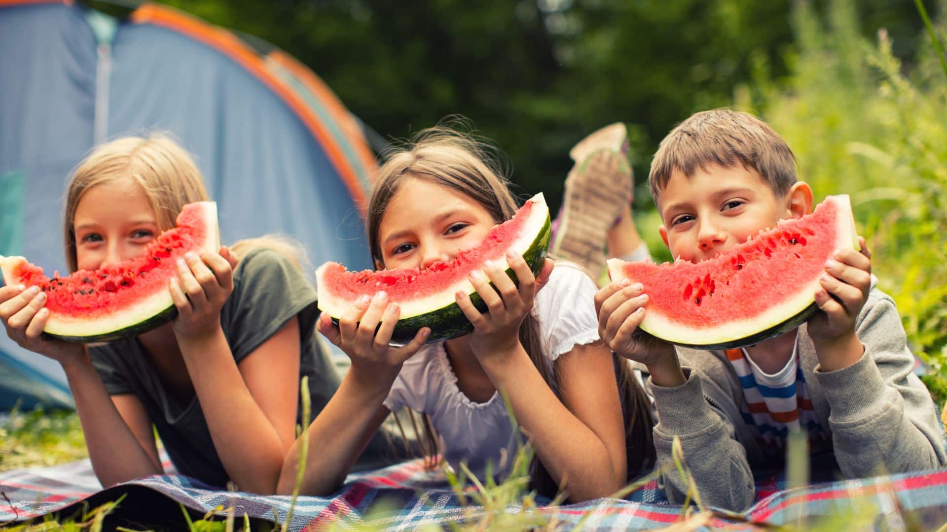Three children eating watermelon slices outdoors in a sunny garden setting.
