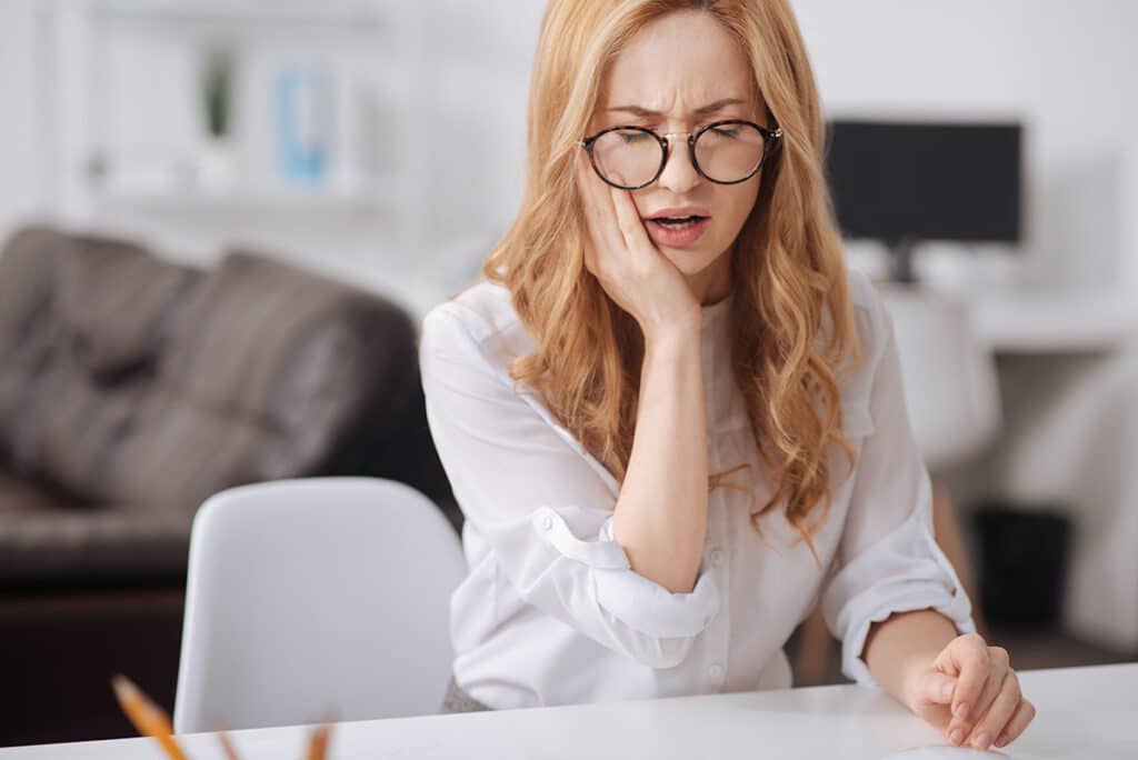 Woman wearing glasses at desk in modern office, smiling while working on laptop.