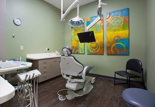 Modern dental operatory with chair, equipment, colorful artwork, and patient waiting area in clinical setting.