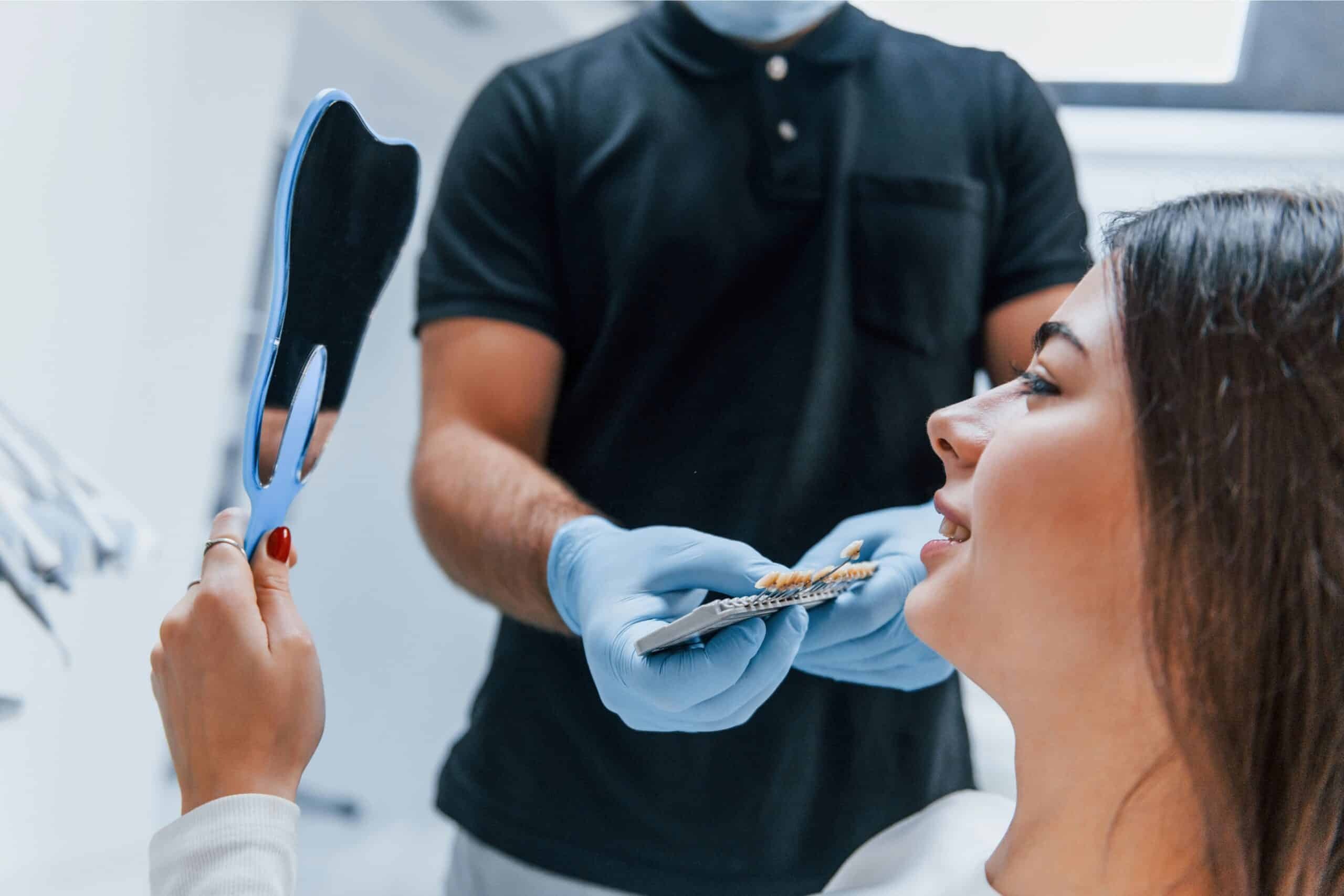 Woman viewing her smile in mirror while dentist holds dental shade guide during cosmetic dental consultation.